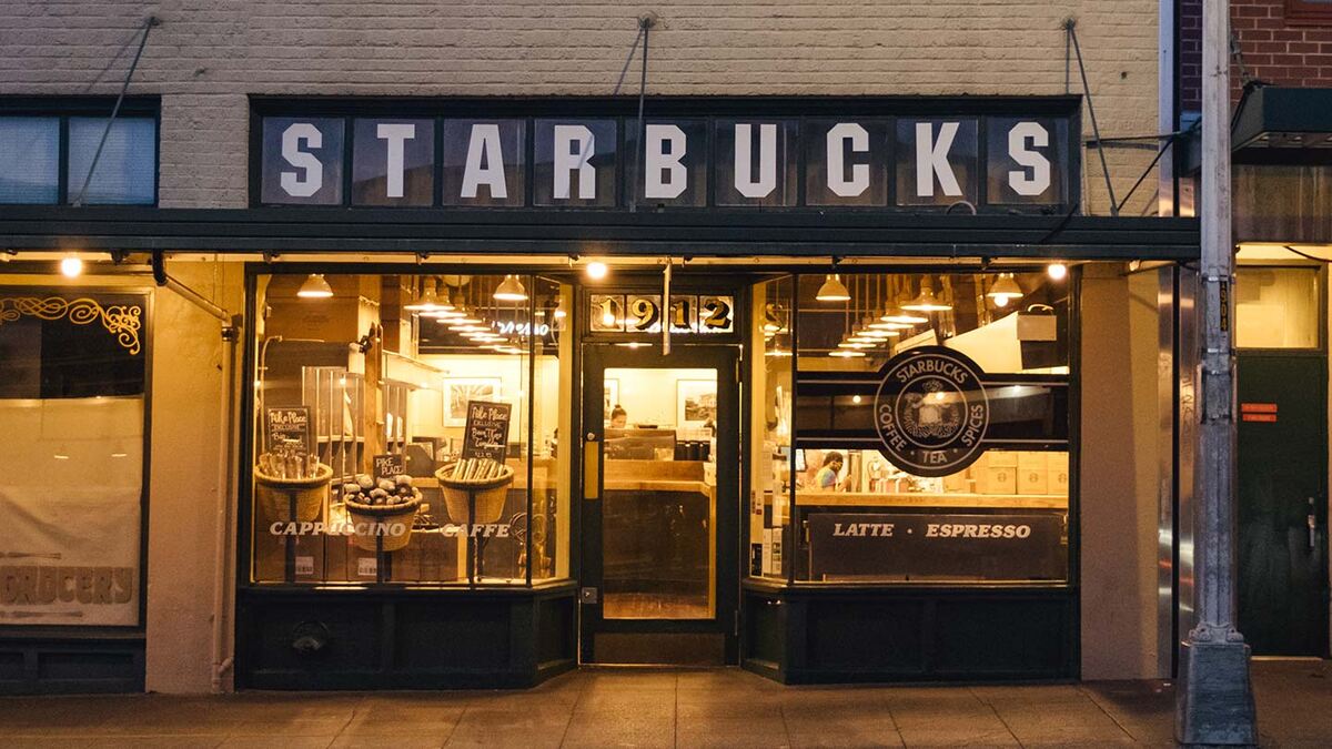 Original Starbucks coffee shop storefront with vintage signage, large display windows, and warm interior lighting at dusk