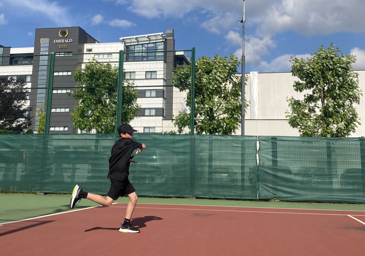 Rion dressed in black shorts, t-shirt, and sneakers hitting a forehand on a tennis court