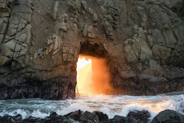 The image shows waves crashing against a rocky shore, with sunlight streaming dramatically through a large, natural archway in the rock formation. The golden light creates a striking contrast against the dark, rugged stone, casting a warm glow onto the mist and spray from the ocean.