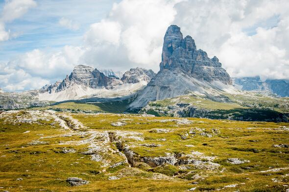 A scenic photograph of a mountainous landscape, prominently featuring the Dolomites in northeastern Italy. The towering rock formation in the center is characteristic of this mountain range, with its steep vertical walls and rugged peaks. In the foreground, there is a grassy alpine plateau with scattered rocks and a visible path or trench leading toward the mountains. The sky is partly cloudy, casting dynamic light and shadow over the scene.