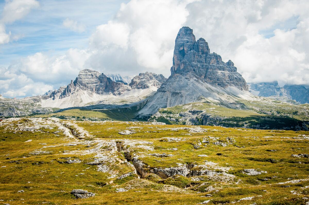 A scenic photograph of a mountainous landscape, prominently featuring the Dolomites in northeastern Italy. The towering rock formation in the center is characteristic of this mountain range, with its steep vertical walls and rugged peaks. In the foreground, there is a grassy alpine plateau with scattered rocks and a visible path or trench leading toward the mountains. The sky is partly cloudy, casting dynamic light and shadow over the scene.